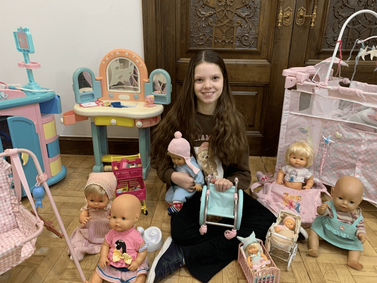 Ein Mädchen lächelt auf dem Boden sitzend, umgeben von Puppen und Spielzeug, und hält einen Spielzeugrollstuhl.
