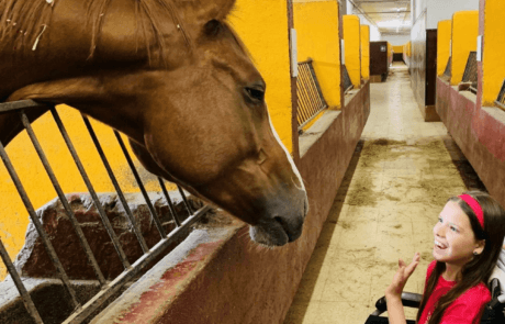 Carolina begrüßt in ihrem Rollstuhl ein Pferd, das den Kopf aus dem Stall streckt.