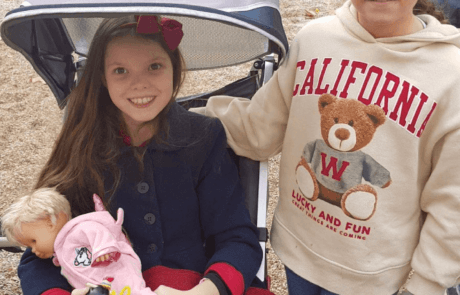 Carolina, in ihrem angepassten Stuhl und mit einer Puppe im Arm, im Park neben ihrer besten Freundin Patricia.