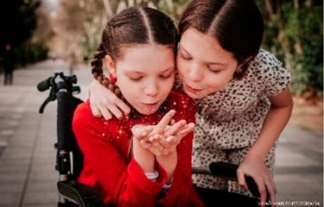 Carolina in ihrem Rollstuhl und ihre Schwester Isabel pusten gemeinsam eine Handvoll Glitzer.