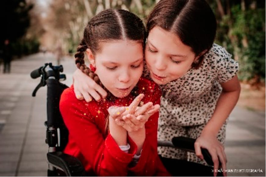 Carolina in ihrem Rollstuhl und ihre Schwester Isabel pusten gemeinsam eine Handvoll Glitzer.