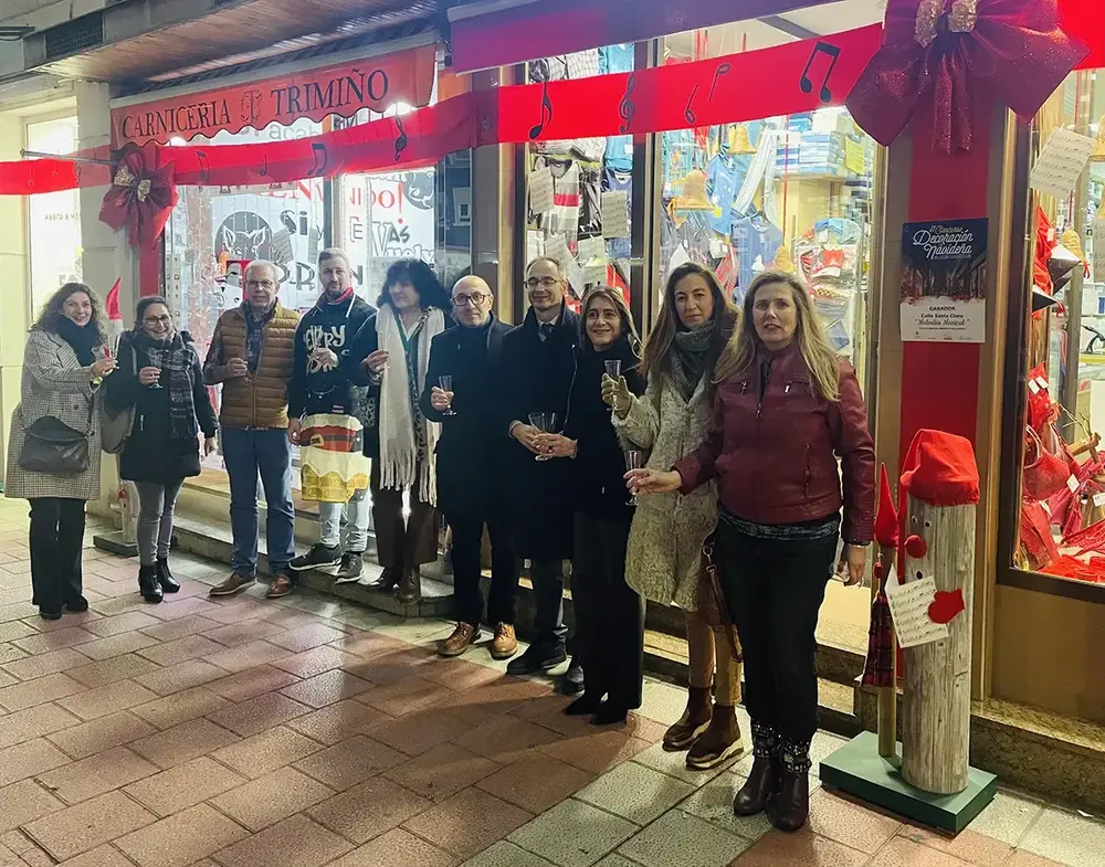 Eine Gruppe stößt mit Gläsern vor einem mit roten Schleifen und Notenzeichen geschmückten Geschäft an, neben dem Plakat des Weihnachtswettbewerbs.