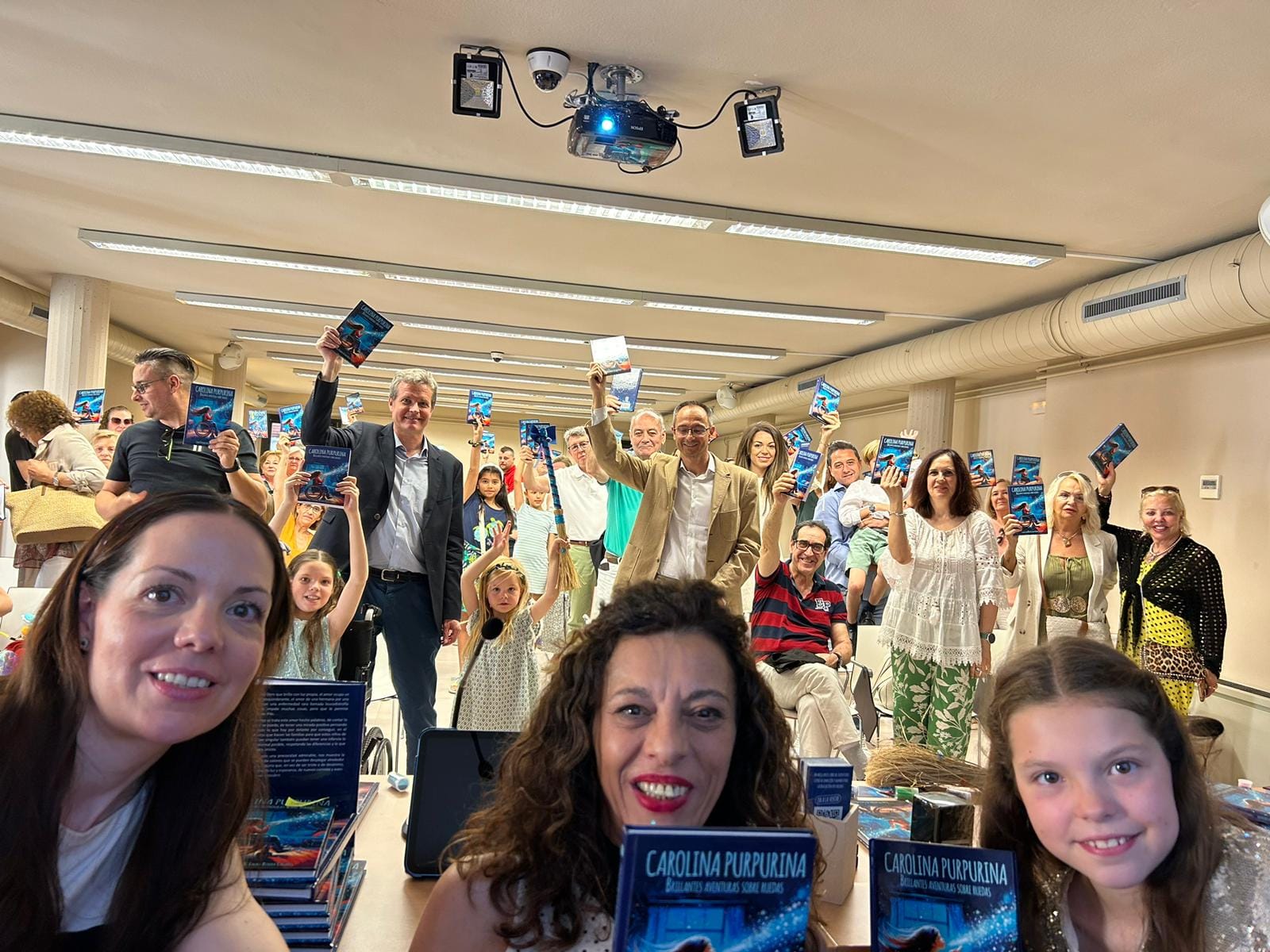 Selfie bei der Buchvorstellung: Das Publikum hebt Exemplare von „Carolina Purpurina“ über einem Tisch voller Bücher.