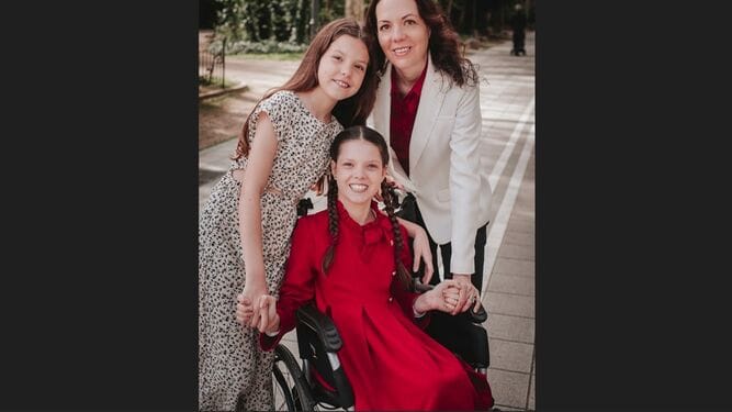 Porträt im Freien: Carolina, in Rot in ihrem Rollstuhl, hält ihre Schwester und ihre Mutter an der Hand, die sie umarmen.