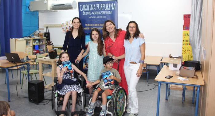 Gruppenfoto in einem Klassenzimmer vor dem Roll-up des Buches: drei Frauen, ein Mädchen und zwei Kinder im Rollstuhl mit Exemplaren.