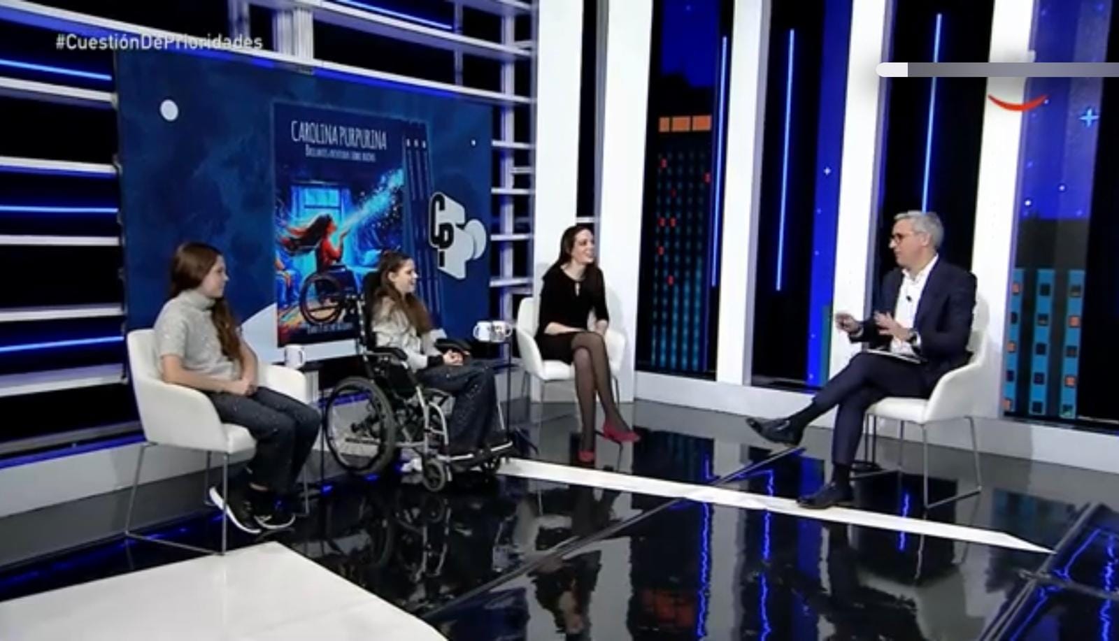 Fernsehstudio: zwei Mädchen, eines im Rollstuhl, und eine Frau unterhalten sich mit dem Moderator vor der Titelseite des Buches.