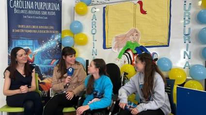 Interview von COPE in einer Schule: eine Journalistin mit Mikrofon neben Carolina und zwei weiteren Mädchen, vor dem Roll-up des Buches.