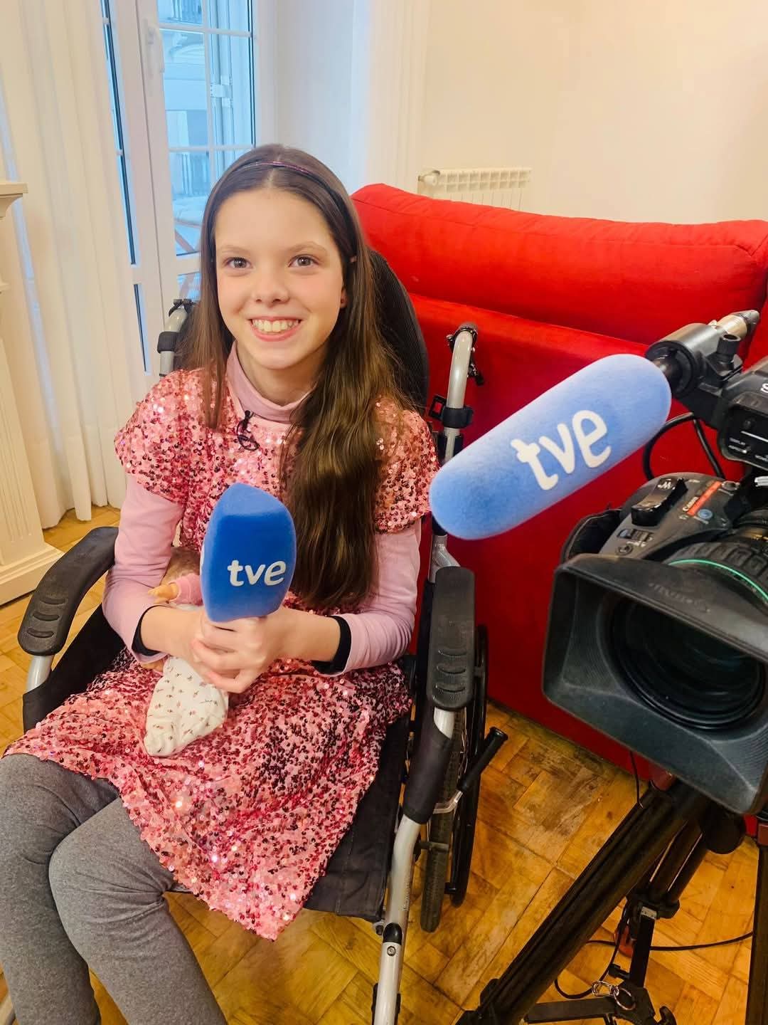 Carolina hält in ihrem Rollstuhl und in rosa Paillettenkleid ein Mikrofon von TVE vor der Kamera.