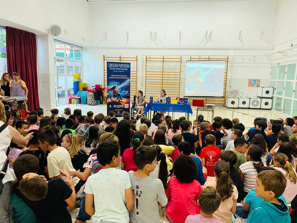 Dutzende auf dem Boden sitzende Schulkinder hören zwei Mädchen am Tisch zu, eines im Rollstuhl, neben dem Roll-up des Buches.