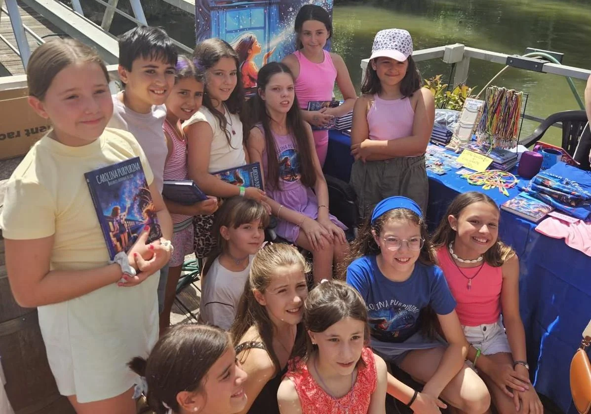 Eine Gruppe von Mädchen posiert neben dem Verkaufsstand am Flussufer; mehrere halten das Buch „Carolina Purpurina“.