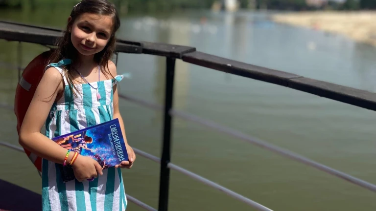 Ein Mädchen mit gestreiftem Kleid und Rucksack hält ein Exemplar von „Carolina Purpurina“ am Geländer an einem Fluss.