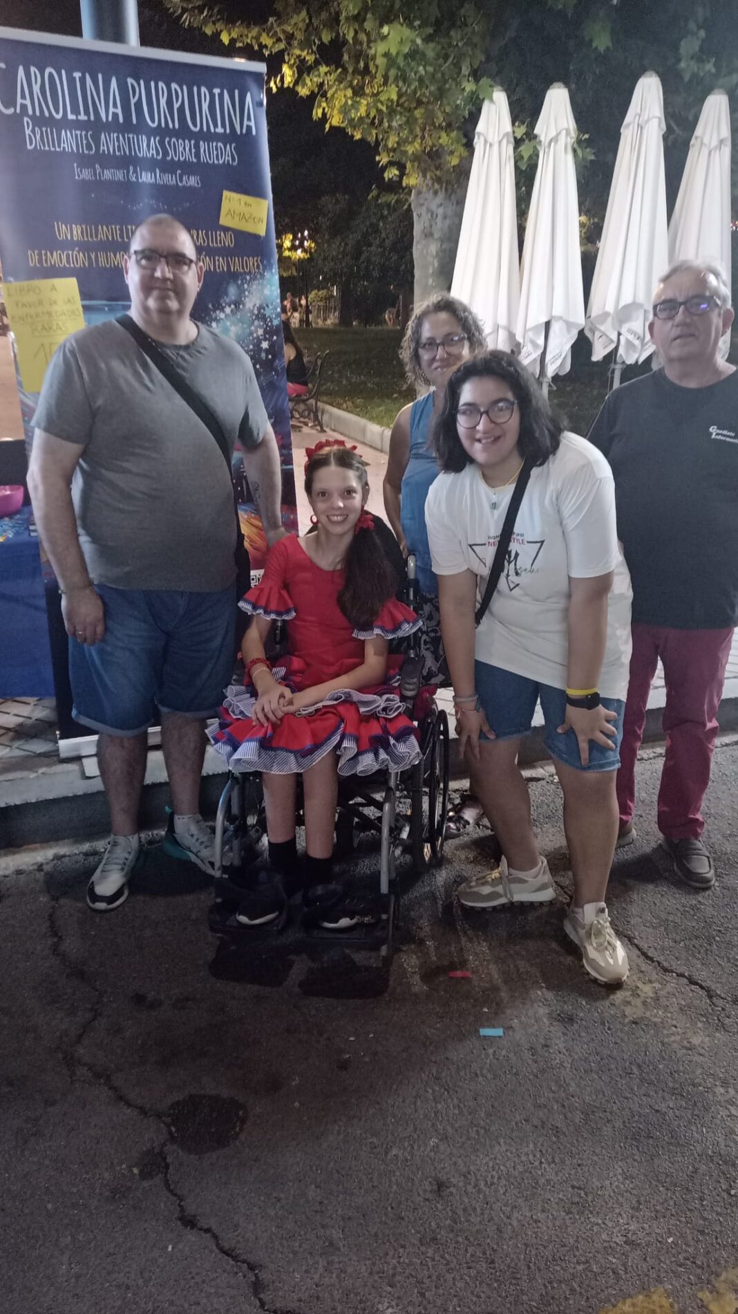 Nachts auf dem Volksfest posiert ein Mädchen im Flamencokleid im Rollstuhl mit mehreren Personen vor dem Roll-up des Buches.