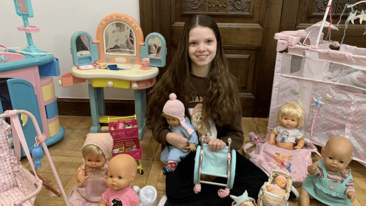 Isabel sitzt zwischen ihren Puppen auf dem Boden und hält einen Spielzeugrollstuhl.
