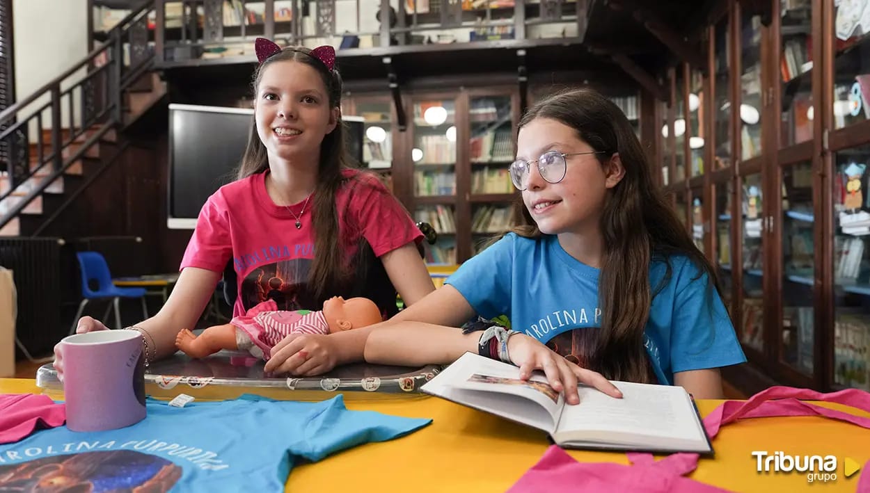 Carolina und Isabel in einer Bibliothek, mit dem aufgeschlagenen Buch, einer Puppe und T-Shirts von Carolina Purpurina.