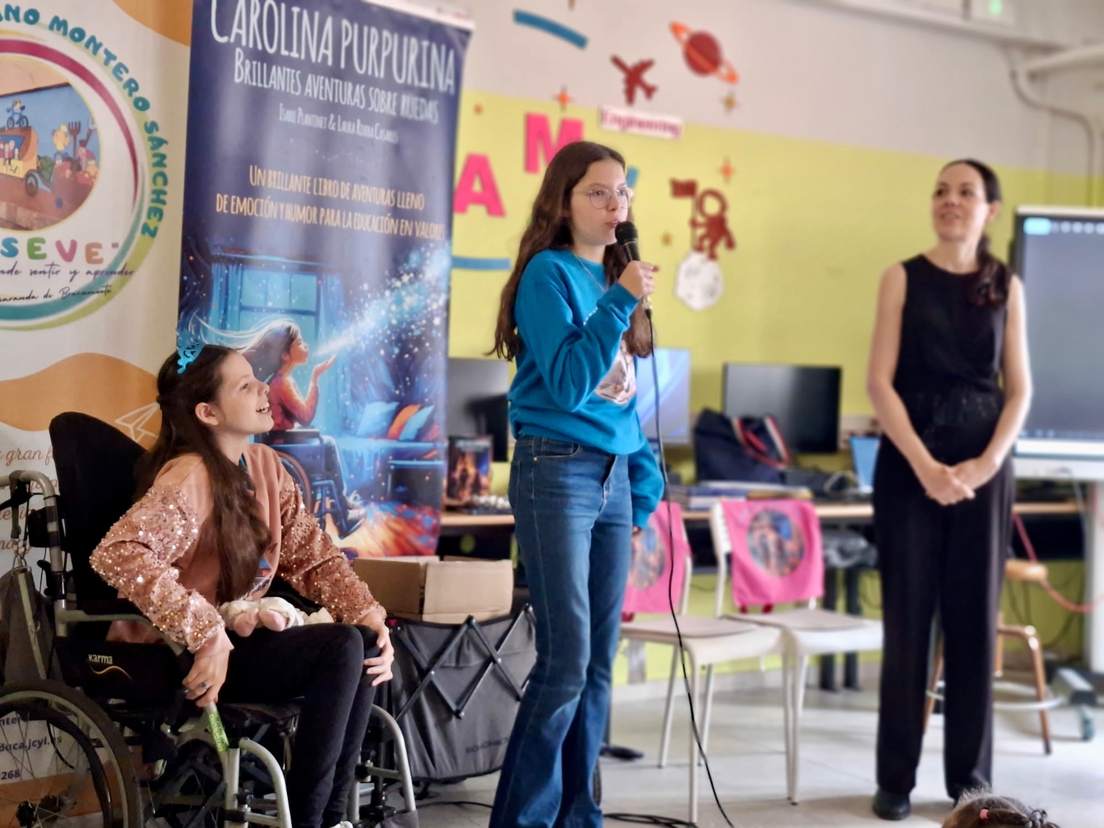 Isabel spricht während der Schulveranstaltung ins Mikrofon, mit Carolina und Laura an ihrer Seite.