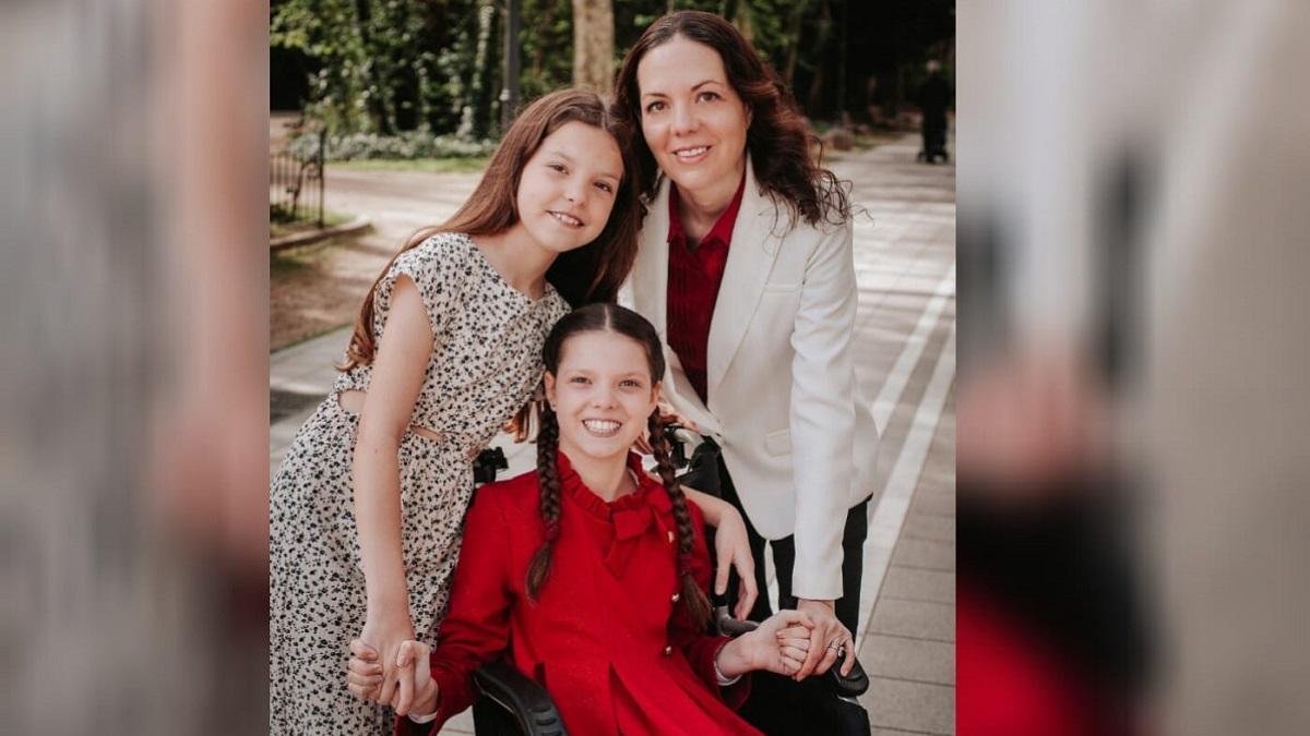 Carolina in rotem Kleid und mit Zöpfen lächelt in ihrem Rollstuhl, neben einem Mädchen im gemusterten Kleid und einer Frau in weißer Jacke.