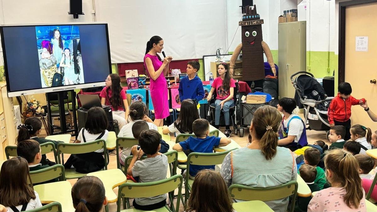Eine Frau spricht vor den sitzenden Schülern ins Mikrofon; neben ihr einige Kinder und Carolina im Rollstuhl an einem Tisch mit Büchern.