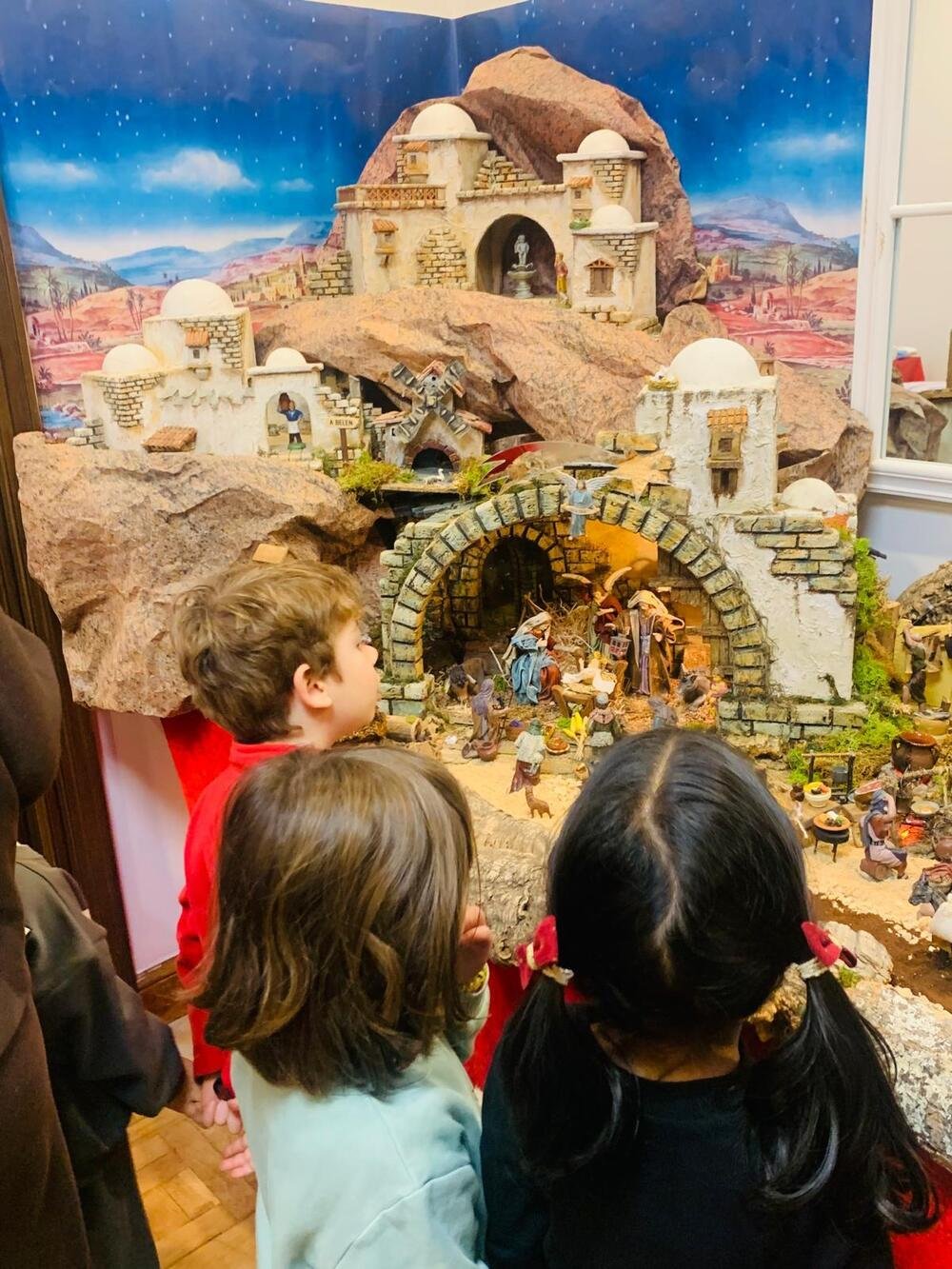 Drei Kinder, von hinten gesehen, betrachten die Krippe: weiße Häuser, ein Stall aus Stein mit der Geburtsszene und ein Sternenhimmel.