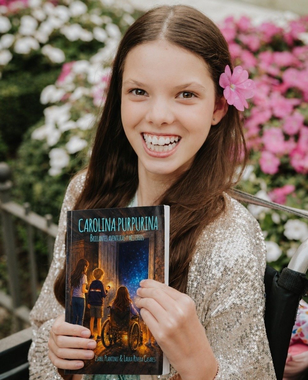 Ein lächelndes Mädchen mit rosa Blüte im Haar hält das Buch „Carolina Purpurina. Brillantes aventuras y misterios“.
