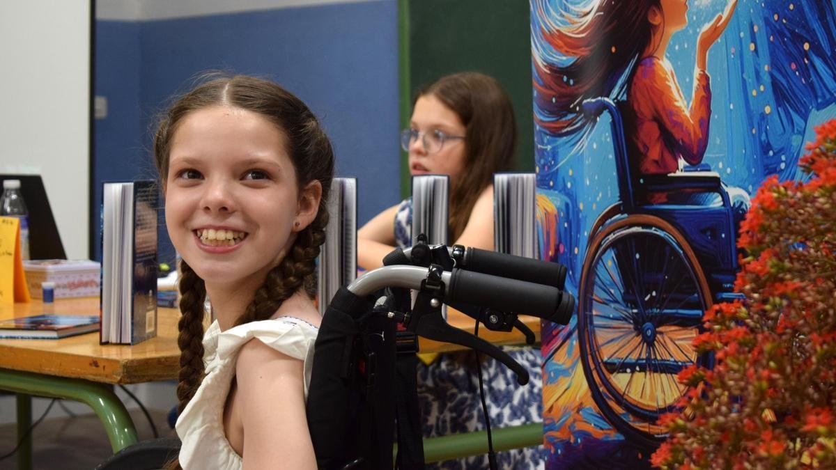 Buchvorstellung: Carolina lächelt in ihrem Rollstuhl neben dem Banner mit dem Cover; dahinter sitzt ein Mädchen am Büchertisch.