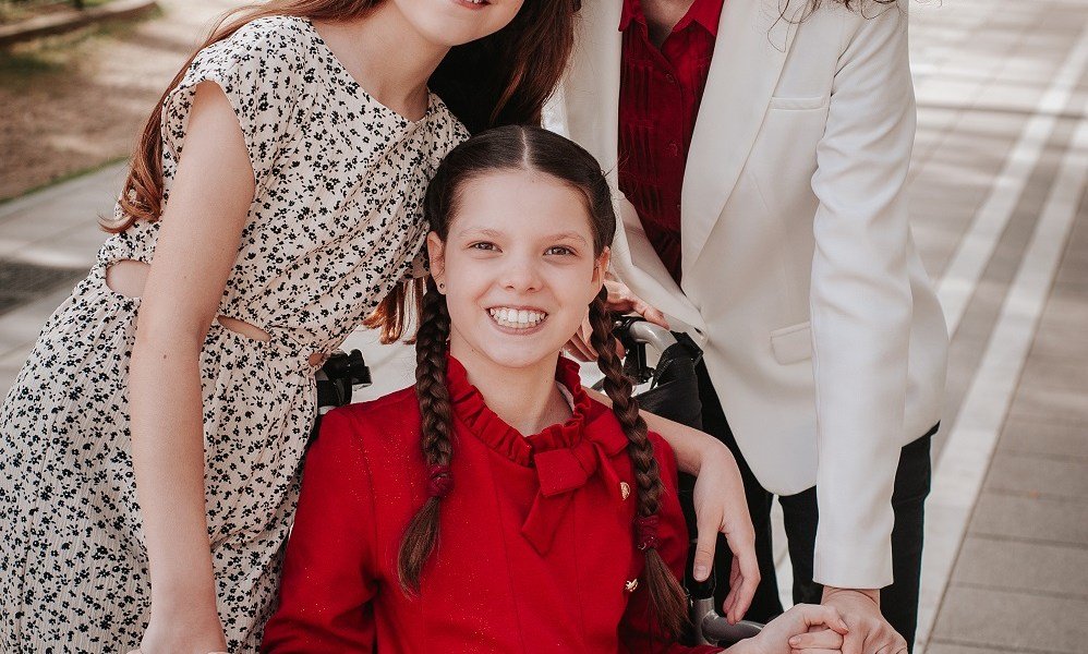 Carolina in rotem Kleid und mit Zöpfen lächelt in ihrem Rollstuhl, umarmt von ihrer Familie, deren Gesichter angeschnitten sind.