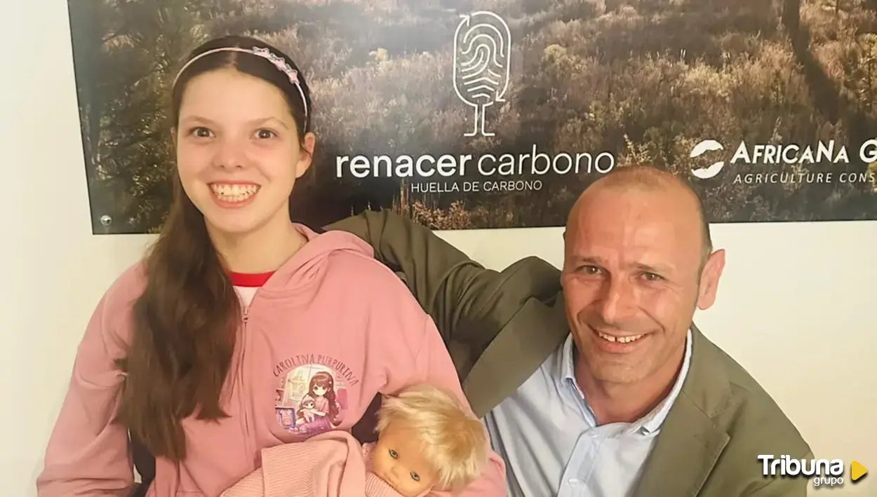 Ein Mädchen im rosa Carolina-Purpurina-Pulli, eine Puppe im Arm, posiert mit einem Mann vor dem Banner von Renacer Carbono.